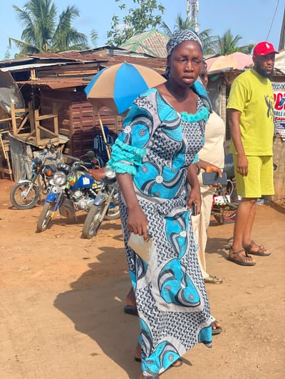 A woman in a blue and white patterned dress and headwrap is photographed at eye level in what appears to be a roadside setting in Ipota, Nigeria. She is holding a small, clear plastic bag in her left hand and has a somber expression. Behind her, partially obscured by a blue and brown umbrella, is a dilapidated wooden structure with a corrugated metal roof. Two motorcycles are parked in front of the structure. A man wearing a yellow shirt and shorts, and a red baseball cap is standing in the background. Another person is lying on a light colored mattress or blanket between the woman and the man. The ground is dirt, and there are palm trees in the background.