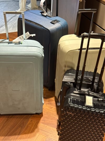 The image shows a collection of four suitcases on a wooden floor, likely indoors. In the foreground, a light blue suitcase is prominently displayed, with its telescopic handle extended. Behind it and to its right, a dark blue suitcase with the "Samsonite" logo is visible. Further to the right, a light beige suitcase is partially obscured. In the very foreground, a black suitcase with a textured geometric pattern and the "STTELLI" logo is present, also with its handle extended. Luggage tags are attached to some of the suitcases. The lighting suggests an indoor environment, possibly a hotel lobby or a personal residence. The scene is devoid of any people or animals. The location in Zapopan, Mexico is not discernible from the visual information provided.