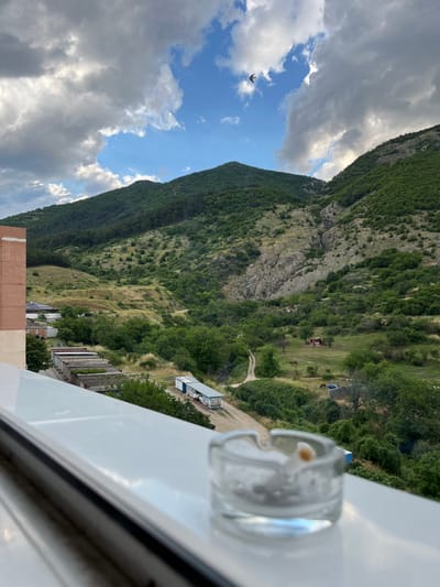 The image shows a view from a balcony or window overlooking a lush green valley with rolling hills and mountains in the background. In the foreground, on the ledge, sits a glass ashtray with a cigarette butt in it. The valley is dotted with trees, and a dirt road winds through it. A few horses can be seen grazing in a field. In the middle ground, there are some buildings, including what appears to be a small industrial structure and a few mobile homes. The sky is partly cloudy, with patches of blue sky visible. The overall impression is one of a rural or semi-rural setting, possibly in the outskirts of a town or city.
The view from this location in Sopot, Bulgaria, offers a scenic panorama of the surrounding natural landscape. The buildings in the foreground suggest a residential area, perhaps an apartment complex or a guesthouse, with balconies providing access to the view. The mountains in the distance are characteristic of the Balkan Mountains, which Sopot is nestled within. The presence of the ashtray indicates that someone was recently smoking on the balcony.