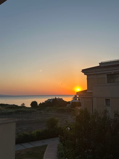 The image depicts a coastal sunrise scene in Chernomorets, Bulgaria. In the foreground, a portion of a beige building with a red-tiled roof is visible on the right side. To the left and slightly lower, another beige building is partially seen. The middle ground features a dark, fallow field bordered by a fence, with scattered green bushes and trees. Beyond the field, the calm sea stretches to the horizon, reflecting the warm hues of the rising sun. Several buildings, some appearing modern with white facades, are situated along the coastline. The sky transitions from a pale blue at the top to a vibrant orange and yellow near the horizon, indicating the early stages of sunrise. Two birds are captured in flight against the colorful sky. The overall atmosphere is serene and tranquil, capturing the beauty of a coastal morning.