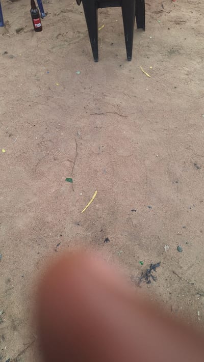 The image shows a sandy ground with several objects scattered across it, in what appears to be an outdoor area in Kporva, Nigeria. In the upper left corner, a brown bottle, likely a beer bottle, stands upright. To its right, the legs of a black plastic chair are visible, suggesting a seating area. The ground is a light brown sand, with various small debris, including what appear to be dry leaves and twigs, interspersed. Some of these pieces are yellow, and one small green object is also visible. The image also features a blurred section in the lower part, which appears to be a person's hand or finger, indicating that a person was likely taking the photograph. The scene suggests a casual outdoor setting, possibly a yard or a gathering place, with the presence of a drink and seating.