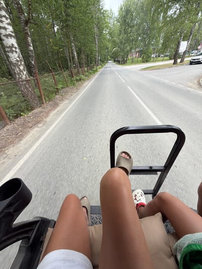 The image shows a view from the back of a vehicle, possibly a golf cart or similar recreational vehicle, driving down a paved road lined with trees. In the foreground, the legs and feet of two people are visible. One person is wearing tan slides and the other is wearing white clogs with a cartoon character design. The road has white lane markings and a white shoulder line. On the left side of the road, there is a metal fence and dense foliage. On the right side of the road, more trees and some buildings can be seen in the distance. The sky is overcast. The location is identified as Morozovo, Russia.