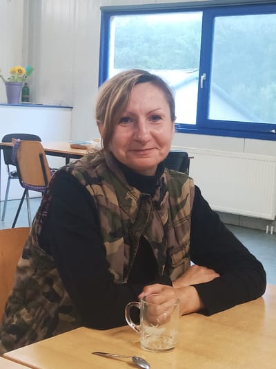 A medium shot, portrait-oriented photograph shows a Caucasian woman seated at a light brown wooden table. The woman is in the foreground, facing the camera, with her arms crossed and resting on the table. She has blonde hair, styled with some bangs swept to the side, and she is wearing a black turtleneck under a camouflage vest. A clear glass mug, with some white residue at the bottom, sits on the table to her right, along with a spoon.
The background of the image depicts a classroom or communal space. To the left, a light brown table and orange chair with a purple seat cover are visible. In the far left background, there is a shelf with a purple vase containing yellow flowers and a green bottle. Directly behind the woman, a large window with blue framing overlooks an outdoor scene of trees and buildings, suggesting it is daytime. A white radiator runs along the wall beneath the window. The floor is a neutral gray color. The scene appears to be an indoor common area, possibly in Sexbierum, Netherlands, given the context.