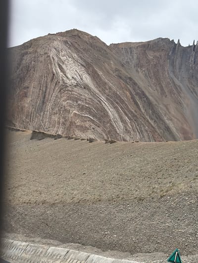 The image shows a mountainous landscape with distinctive geological formations. The mountain face exhibits striking patterns of folded rock strata, with alternating bands of light and dark colors, suggesting immense geological pressure over time. The foreground is a gravelly slope leading up to the base of the mountain. In the lower right corner of the image, a triangular green and white road sign is partially visible. The sky is overcast with a light gray hue. The overall scene depicts a remote, natural environment, likely a mountainous region. The location is identified as Stongde, India.