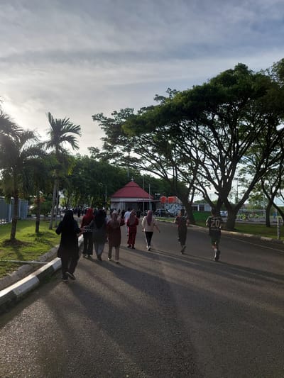 The image depicts a group of people walking and running on an asphalt road in Banda Aceh, Indonesia. The scene is characterized by a bright, partly cloudy sky and lush green trees lining the road. On the left side of the road, there is a grassy area with palm trees and a white curb with black markings. In the background, a building with a red roof and white walls is visible, along with more trees and a sports field. The people are dressed in casual clothing, with some wearing hijabs, suggesting a warm climate. The lighting suggests it is either early morning or late afternoon, with long shadows cast by the trees and people on the road. The overall atmosphere appears to be one of casual outdoor activity.