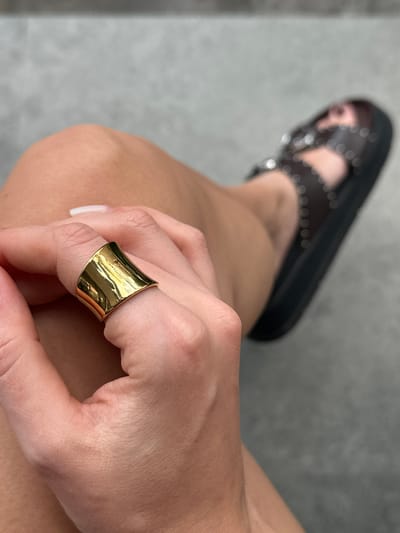 A person's hand, adorned with a large, polished gold ring on the index finger, rests on their leg. The ring has a flared, curved shape. The background is blurry but reveals a brown sandal with silver studs on the person's foot, suggesting they are seated. The setting is indoors with a gray floor. This image does not contain enough information to confirm or deny the location in Kerobokan Kelod, Indonesia.