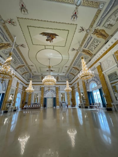 The image shows the interior of a grand hall, likely a ballroom or reception room, within the Mikhailovsky Palace in Strelna, Russia. The hall is ornately decorated with gilded chandeliers, intricate ceiling frescoes, and marble columns. The ceiling features detailed paintings and decorative moldings. Several large, multi-tiered chandeliers hang from the ceiling, illuminating the room with a warm glow. The floor is highly polished, reflecting the chandeliers and architectural details. In the background, there are large arched windows draped with blue curtains, and a set of double doors. Rows of chairs are visible along the walls, suggesting the space is used for events. The overall impression is one of opulence and historical grandeur.
