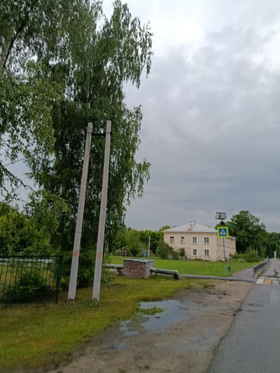 This media file depicts an outdoor scene in Vereya, Russia. In the foreground, two weathered concrete poles stand amidst lush green grass. A dark green metal fence runs along the left side of the frame, partially obscured by dense foliage. The ground in the immediate foreground is a mix of wet asphalt and grass, with several puddles indicating recent rain.
In the midground, a light-colored building with a light grey roof is visible. The building appears to be a two-story structure with multiple windows. In front of the building, a pedestrian crossing sign with a blue and yellow triangle depicting a person walking is prominently displayed. To the left of the building, a brick structure and a horizontal pipe are present.
The background is dominated by a canopy of green trees and a cloudy, overcast sky. The overall atmosphere suggests a subdued or overcast day, likely in the late afternoon or early evening, given the soft lighting. There are no people visible in the scene. The weather appears to be humid or recently rained. The presence of the pedestrian crossing sign and the building suggest this might be a residential or semi-rural area. There is no visible text in the image.