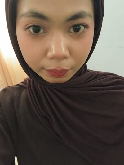 A close-up, frontal shot captures a young woman wearing a dark brown hijab and top, likely indoors. She is looking directly at the camera with a neutral expression. Her makeup includes defined eyebrows, mascara, and a touch of blush on her cheeks. Her lips are painted with a reddish lipstick, and a subtle highlight is visible on her nose and forehead, suggesting good lighting conditions. The background is a plain, off-white wall with a soft yellow curtain to the left. The lighting appears to be artificial. The image is a selfie, taken at eye level with the subject. The subject's identity and the specific location within Watampone, Indonesia, are not discernible from the visual information alone.