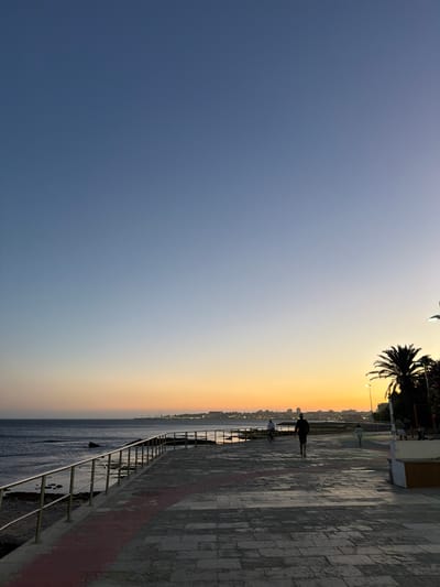 This image depicts a sunset scene along the coast in Cascais, Portugal. The sky transitions from a clear blue at the top to hues of orange and yellow near the horizon, indicating the approach of evening. A paved promenade runs parallel to the sea, bordered by a metal railing. Several individuals are visible on the promenade: one person is riding a bicycle, another is walking towards the viewer, and a third, a child, is running away from the viewer. In the background, a line of buildings and palm trees suggest an urban or resort area. The sea is calm, with gentle waves lapping against the shore. The overall atmosphere is serene and depicts a typical evening scene in a coastal town.
