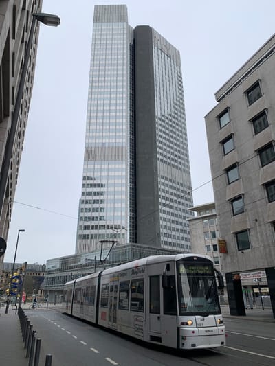 The image shows a white tram traveling on a street in Frankfurt am Main, Germany. The tram is traveling to the right. A tall glass and steel office building is visible in the background. The office building is located on the left side of the image. There is a tall, gray concrete building on the right side of the image. The tram is near the corner of the buildings. The street is empty, except for the tram. The tram is marked with the number "4" and the words "Bornheim-Eisentrautplatz". The tram is labeled with advertisements for "Main Viertel". The tram is travelling on a paved street. There are trees and sidewalks on the left and right sides of the street. There are no people visible in the photo. The image was taken in the city of Frankfurt am Main, Germany, at coordinates (latitude: 50.11, longitude: 8.675). The sky is cloudy.