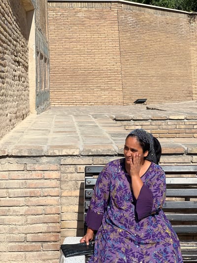 In Samarkand, Uzbekistan, a woman sits on a dark wooden bench against a backdrop of weathered brick walls and paved stone steps. She wears a vibrant purple dress adorned with a floral pattern and a matching headscarf with intricate beaded embellishments. Her right hand is raised to her cheek, suggesting a thoughtful or perhaps weary pose. The lighting indicates it is daytime, with shadows cast on the ground and the wall. The architecture suggests an ancient or historical setting, possibly a courtyard or a historical site within the city.