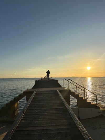 A person is walking on a wooden pier extending into the ocean at sunset. The sky is clear with a gradient from light blue to orange. The sun is setting on the horizon, casting a bright reflection on the water. Islands are visible in the distance. Another person is also on the pier further away. A surfboard rests on the sand near the base of the pier. The pier is equipped with stairs and handrails. The scene captures a tranquil evening in Baan Taling Ngam, Thailand.