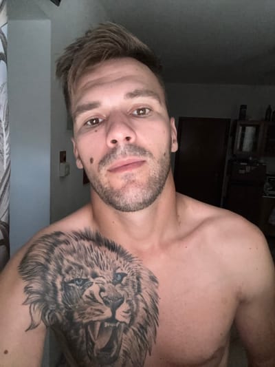This is a close-up, sideways selfie of a man with a lion tattoo on his right shoulder. The man has short, light brown hair, brown eyes, and a light beard. He is looking directly at the camera with a neutral expression. The background is dimly lit, showing a wall and some shelves with objects, suggesting an indoor setting. The lighting indicates it might be evening or a dark room. There is no visible text in the image. The scene appears to be a personal snapshot taken in a private space.