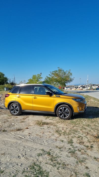 In Limani Litochorou, Greece, a yellow Suzuki Vitara SUV is parked on a sandy and grassy area next to a paved road. The vehicle is positioned facing right, with its front right wheel and rear left wheel visible. The SUV has a black roof and tinted windows. Behind the car, a clear blue sky stretches above a scene of a harbor with several boats docked, some with masts. Trees and some structures are also visible along the waterfront. The ground in the foreground shows tire tracks in the sand. There are no people visible in the image.