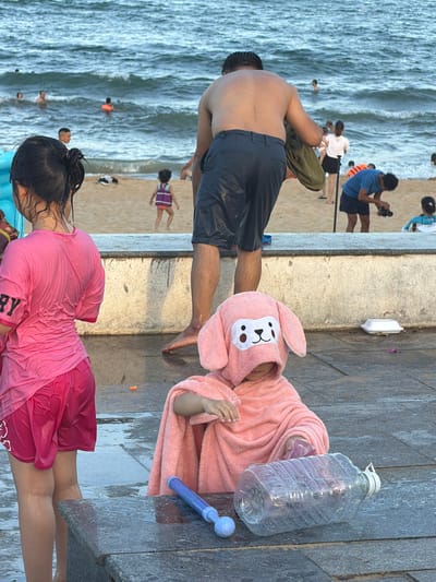This media file captures a lively scene at a beach in South Quy Nhon Ward, Vietnam. The image features several individuals, including a man in dark shorts standing on a paved walkway, his back to the camera, with a towel draped over his shoulder. Nearby, a child is nestled in a pink, fuzzy bathrobe adorned with a bunny face.
In the background, the ocean is visible with waves crashing onto the shore, and several people are in the water. The sandy beach also has a few individuals, including a child in a purple and white dress.
On the paved area, a large, clear plastic water bottle lies on its side, and a blue water gun is positioned next to it. Another person, a young woman, is in the foreground, wearing a wet pink t-shirt and red shorts. Her hair is dark and wet, pulled back from her face.
The scene suggests a warm, sunny day, likely during daylight hours, given the brightness and the beach setting. The presence of water guns and the wet clothing indicate a day of play and cooling off in the water. The overall atmosphere appears relaxed and recreational.