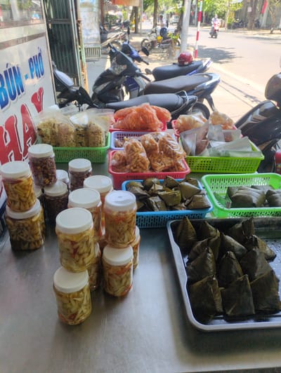 The image shows a street food vendor's setup in Tuy Hòa, Vietnam. Several containers and baskets hold various food items: rice dumplings wrapped in green leaves, crispy snacks in plastic bags, pickled vegetables in clear plastic jars, and nuts in clear plastic jars. The vendor's cart has the name of the food "Bún-Phở" and some motorbikes are parked on the side of the road behind the cart, suggesting a busy street.