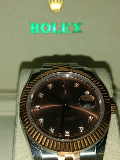 A close-up shot showcases a Rolex watch, likely a Datejust model, with a dark dial and diamond hour markers. The bezel is made of fluted gold, and the watch has a two-tone bracelet, suggesting a combination of steel and gold. The watch is presented in a box, with the brand's logo, "ROLEX", illuminated in green above it. The lighting suggests an indoor setting, possibly a retail display or a personal collection. No people are visible in the image. The overall impression is one of luxury and fine craftsmanship. There is no contextual information about the time of day, weather, or specific location beyond "Ashkelon, Israel" which is not visually represented in the image. The text "ROLEX", "OYSTER PERPETUAL", "DATEJUST", "SUPERLATIVE CHRONOMETER", and "OFFICIALLY CERTIFIED" are visible on the watch face and bezel.