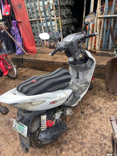 The image shows a white scooter lying on its side on a dirt ground. The scooter is partially damaged, with some of its body panels detached. A person's feet, wearing black flip-flops, are visible next to the scooter. In the background, there are stacked items wrapped in plastic bags and a metal shelving unit. A red tarp and some purple fabric are also visible. A license plate with the text "ENUGU", "FOR CITY STATE", and "ENZ-671 DT" is attached to the scooter. The overall scene appears to be outdoors, possibly in a market or a storage area. The lighting suggests it is daytime, but the weather is unclear. The image is taken from a high angle, looking down at the scooter and its surroundings.