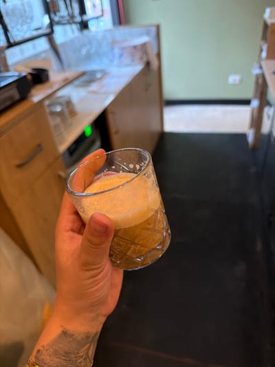 A person's hand is holding a clear glass with a light-colored frothy liquid, possibly a beverage, inside. The glass has a diamond-shaped pattern on its exterior. The hand has tattoos on the wrist and forearm, and is wearing a gold bracelet. The scene appears to be indoors, likely in a cafe or bar, with wooden surfaces and a dark wall in the background. The lighting suggests it could be daytime or evening. The image is slightly tilted.