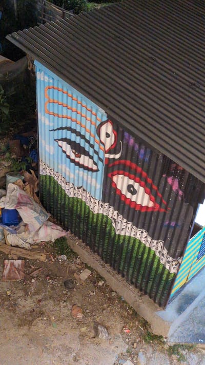 This image, taken in Dharamkot, India, shows an elevated view of a corrugated metal structure. The structure's side wall is composed of vertically oriented corrugated galvanized iron sheets, entirely covered by a painted mural. The mural is vertically split into two distinct background themes. The left section features a light blue sky background with white cloud-like formations and horizontal orange wavy lines at the top. The right section displays a dark grey background with diffused pink and purple elements, suggesting a cosmic theme. Both sections share a lower horizontal band featuring vertical green stripes with a jagged white and black patterned border running along the top edge of the green section. Within the left, sky-themed portion, a large, horizontally oriented ovoid shape with black and white concentric patterns and a central black dot is depicted. In the right, cosmic-themed portion, a similar large ovoid shape is present, featuring red and white concentric patterns with a central black dot. A smaller, irregular white shape with a black dot is positioned near the top center where the two mural sections meet.

The roof of the structure is made of dark grey corrugated galvanized iron sheets, oriented horizontally and sloping downwards from left to right.

The ground surrounding the structure is unpaved, consisting of compacted dirt, various sizes of rocks, and scattered debris. Immediately to the left of the painted wall, several crumpled white sacks or bags, a blue plastic bucket, and fragments of wood are visible. A narrow, light-colored concrete or cement strip forms the base of the painted wall. Sparse green vegetation is present along this base and in other areas of the unpaved ground. To the far right, a section of another structure or wall is visible, painted with diagonal yellow and blue stripes. The background beyond the structure shows darker green foliage. No individuals are present in the scene.