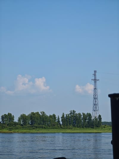 The image shows a wide, calm river in the foreground, with gentle ripples on its surface. On the opposite bank, a lush green treeline stretches across the horizon, dotted with a variety of trees. Prominently visible in the middle ground, to the right of the trees, is a tall, dark, lattice-work power transmission tower, its metal structure silhouetted against the bright blue sky. A few wispy white clouds drift in the sky. In the far distance, beyond the treeline, the landscape appears flat and extends to the horizon. A dark, out-of-focus object, possibly part of a boat or structure, is visible in the extreme right foreground, partially obscuring the view of the river and the landscape. The scene is bathed in natural daylight, suggesting it was taken on a clear day. The location is identified as Verkhnepashino, Russia.