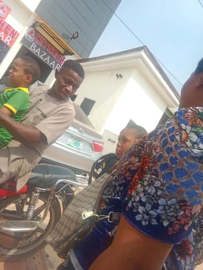 An outdoor scene in Aguata, Nigeria, depicts multiple individuals and vehicles under clear daylight. In the left-center, a man in a tan short-sleeved garment is seated on or straddling a red and chrome motorcycle, holding a young child in a green t-shirt with yellow trim. The man looks slightly right, while the child gazes left. To the immediate right, an individual is partially visible, wearing a blue garment with a multi-colored floral and geometric pattern, with a patterned handbag visible. Another individual is partially discernible behind them, looking left. In the background, a silver Toyota sedan with Nigerian license plate JJT-899KJ is parked. Behind the sedan, a light-colored building displays the partial branding 'Mikano'. To the left of the scene, a commercial building features a red sign with a chicken logo and white text reading "KITCHEN BAZAAR". A modern structure with dark reflective glass is present in the upper left background.