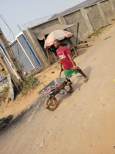 An adult male is depicted pushing a dark green, single-wheeled wheelbarrow filled with numerous red onions on an unpaved, light brown dirt road in Puerto López, Colombia. He is wearing a red t-shirt with white lettering, green shorts, and open-toed sandals. His body is turned slightly, with both hands on the wheelbarrow handles, indicating forward movement. To the immediate left, a concrete utility pole with wires and light vegetation precedes a concrete wall featuring a blue metal gate and topped with barbed wire. Further right in the background, a rudimentary vendor stall is present, covered by a weathered, light-colored umbrella, beneath which various fruits are displayed on a wooden table. Behind this stall, a tall concrete wall topped with coiled barbed wire extends horizontally. The ground is uneven, showing tire tracks and scattered debris. The sky above is clear and light.