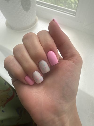A close-up, overhead shot features a woman's left hand displaying a manicure with a pink and white polka dot design on two nails and solid pink on the remaining nails. The hand is positioned over a light-colored, textured surface, possibly a windowsill with a sheer curtain in the background, revealing a glimpse of green foliage outside. The image does not contain any elements that would allow for the determination of its location in Sayanogorsk, Russia.