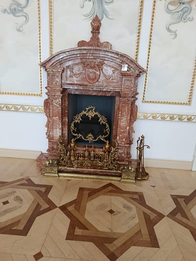 The image shows a gilded fireplace screen in front of a red marble fireplace in a room with ornate walls and a parquet floor. The fireplace is adorned with classical motifs and the screen features intricate scrollwork. To the right of the fireplace, a set of brass fireplace tools stands on a matching stand. The floor is laid with a geometric pattern of light and dark wood. The walls are decorated with white panels featuring gold trim and faint grey artwork. The scene appears to be within a historic building, possibly a palace or museum, located in Kosava, Belarus, likely within the Kosava Palace complex. There are no people visible in the image.
