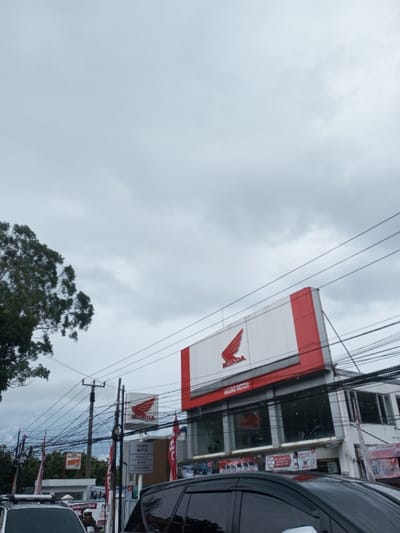 This image depicts a street scene in Cipendawa, Indonesia, observed under an overcast sky with dense grey cloud cover. The primary subject is a multi-story building featuring a Honda motorcycle dealership. A large, rectangular billboard on the upper facade displays the red winged Honda logo and "AGUNG MOTOR" in red text against a white background with red trim. A smaller, square sign with the Honda logo and "AGUNG MOTOR" is visible at ground level to the left. The building's lower floor features large glass display windows, behind which motorcycles are visible. Red banners and flags are present around the building. A utility pole, heavily laden with numerous entangled black electrical and communication wires, stands prominent in the middle-left of the frame. In the foreground, the side of a black minivan or SUV occupies the lower right, and a portion of a white vehicle is visible on the lower left. A single individual wearing a helmet is partially visible on a motorcycle, situated behind the white vehicle. A large tree with dark green foliage is positioned on the far left of the frame.
