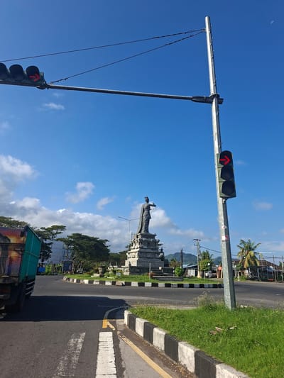 This image depicts a traffic intersection with a statue in the background. In the foreground, a traffic light with a red arrow signal is visible on a metal pole. A truck is partially visible on the left side of the image. The intersection appears to be a roundabout, with grassy medians and decorative borders. In the background, a tall, stone statue of a woman in traditional attire stands on a pedestal. Beyond the statue, buildings and lush green trees can be seen, with mountains in the distance. The sky is a clear blue with a few scattered clouds. This scene is located in the city of Dawan, Indonesia.