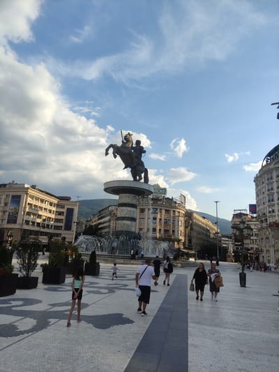 This is a medium-angle, eye-level, outdoor photograph taken in Skopje, North Macedonia, likely in the city center. The primary subject is the Warrior on a Horse statue, which is a prominent monument featuring a bronze equestrian statue atop a circular fountain base. The statue is centrally positioned, with a vast plaza in front of it.
In the plaza, there are several individuals walking and interacting. A young girl wearing a striped tank top and black shorts stands with her back to the camera, facing the fountain. To her right, a young boy in a grey t-shirt and shorts is running away from the camera. In the mid-ground, several adults are walking; one man in a white t-shirt and black shorts carries a white bag, and two women in dark clothing are walking together, one carrying a shopping bag. In the background, more people are visible, walking in various directions.
The environment is urban, with large, ornate buildings lining the plaza. These buildings exhibit classical architectural styles, with multiple stories, balconies, and decorative facades. Signs are visible on some buildings, including "ADMIRAL" and another with an illegible logo. The sky is a vibrant blue with scattered white clouds, and a mountain range is visible in the distance. The plaza is paved with stone tiles, featuring a distinctive pattern. Green planters with trees are strategically placed throughout the plaza. The lighting suggests it is daytime, possibly late afternoon, given the shadows cast on the ground.