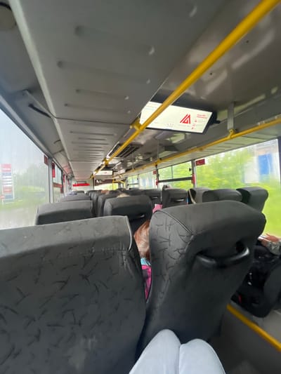 The image is taken from the interior of a bus, looking forward along the aisle. The seats are upholstered in dark gray fabric with a subtle pattern. Yellow handrails run along the ceiling. A digital display above the seats shows route information, including the number "532" and text that appears to be in Russian. Outside the windows, a blurry, green, outdoor environment is visible, suggesting a route passing through a somewhat vegetated area, possibly on the outskirts of a city. In the distance, a red and white sign is visible through the left window, possibly a roadside advertisement. The lighting is overcast, suggesting it may be daytime but not particularly sunny. The image appears to be taken in Krasnokamsk, Russia, based on the prompt.