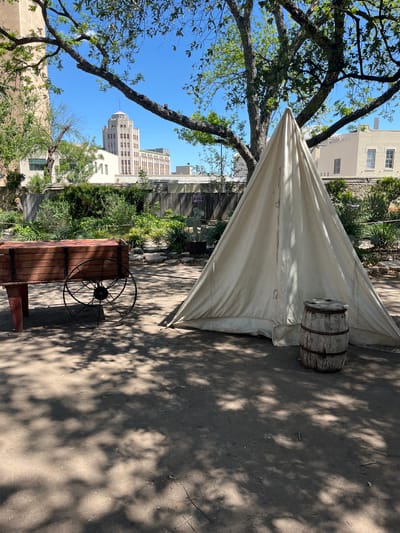 The image shows an outdoor scene in San Antonio, Texas. A white canvas tent stands on the right, with a wooden barrel placed next to it. A wooden cart with a large metal wheel is positioned nearby. The ground is dirt and is covered with shadows. In the background, buildings are visible including the Tower Life Building and other architecture. A tree branch with green leaves is visible in the upper left of the image. The sky is blue and clear.