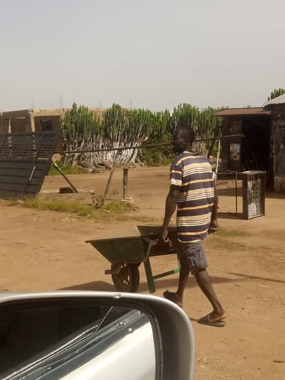 The image shows a man walking with a wheelbarrow on a dirt path in Katon Rikkos, Nigeria. The man is wearing a striped t-shirt and shorts, and he is pushing a green wheelbarrow with a load. In the background, there are several tall, spiky plants and a corrugated metal building. The sky is hazy and light-colored. The foreground shows a portion of a car, likely a side-view mirror and door frame, suggesting the photo was taken from inside a vehicle. The environment appears to be an outdoor area with arid ground and sparse vegetation.