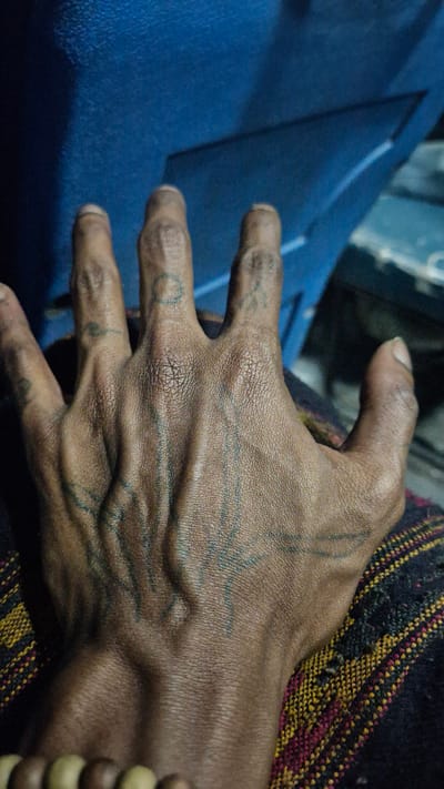 The media file shows a close-up view of a person's hand, exhibiting visible tattoos and prominent veins. The hand is resting on a textured fabric with a pattern of red, yellow, and black threads. To the left of the hand, a bright blue fabric is visible, possibly clothing or upholstery, with a sharp crease indicating a fold. The lighting suggests it is daytime, but no specific weather conditions can be determined. The skin tone of the hand is dark, and the tattoos appear to be abstract line work and possibly a circular design on one of the fingers. The setting does not provide definitive cues for the exact location within Kullu, India, but the presence of traditional-looking textiles might suggest a cultural context. No text is visible in the image. The overall impression is a candid shot focusing on the details of the hand and its adornments, with a hint of the surrounding environment.