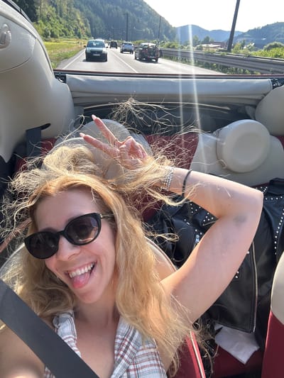 A young woman with blonde hair is taking a selfie in a convertible car on a highway. She is wearing sunglasses and a plaid shirt. Her hair is blowing in the wind, and she is sticking her tongue out and making a peace sign with her hand. The car is red and cream colored. In the background, there are other cars on the highway, as well as trees and mountains. The sun is shining, and there are lens flares in the image. The scene appears to be on a highway passing through a mountainous, wooded area. Based on the visual cues and the prompt, the location is the B33 highway near Hausach, Germany, a route known for its scenic beauty and mountainous terrain.