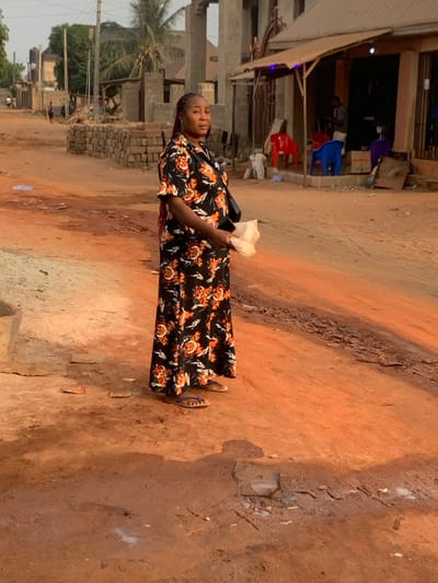 A woman stands on a reddish-brown dirt path in what appears to be a semi-urban or rural setting. She is positioned slightly to the left of the frame's center, looking towards her left with a neutral expression. Her hair is styled in braids. She wears a long, loose-fitting dress featuring a black background with an orange and white floral pattern. On her feet are blue flip-flops. She holds a small, light-colored crumpled object, possibly paper or a food item, in her right hand. A black bag strap is visible over her left shoulder.

The ground is predominantly uneven, reddish-brown dirt, characteristic of laterite soil, with some darker, possibly damp patches or small divots. In the background, to the woman's left, are structures in varying stages of construction or repair, including exposed concrete and brickwork. Palm trees and utility poles are also visible in the distance. To her right, further along the path, is a building with an awning, outside which are a red and a blue plastic chair, suggesting a small shop or gathering area. The overall lighting indicates daylight, likely mid-day, with a bright, somewhat harsh quality. No discernible text is visible in the image. The scene conveys a moment of pause on a well-trodden path in a local community.