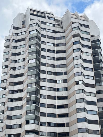 The image shows a tall, modern residential building with a distinctive angular facade. The building is made of white and beige tiled panels, with numerous windows and balconies. The facade features a zig-zag pattern created by alternating projecting and recessed sections. The building is seen from a low angle, emphasizing its height against a cloudy sky with patches of blue. This building is located in Créteil, France, and is part of the city's architectural landscape. There are no people or visible actions occurring in the image; it is a static depiction of the building's exterior.
