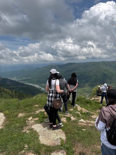 The image depicts a group of people observing a scenic mountain landscape from a high vantage point. The foreground features a grassy, rocky terrain with several individuals standing and sitting. A woman in the front left is wearing a white shirt, a black and white plaid vest, black pants, and a white baseball cap, carrying a brown circular bag. To her right, a man in a grey hoodie and black pants stands with his hands behind his back. Further to the right, a person in a black hooded garment is visible. In the mid-ground, another person is seated on a rock, looking towards the landscape, and a woman in a grey hoodie and jeans is taking a photo with her phone. The background reveals a vast, verdant valley with a river winding through it, surrounded by rolling, forested mountains under a cloudy sky. This location is consistent with views found near Nizhnyaya Teberda, Russia, a region known for its mountainous terrain and natural beauty.