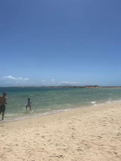 The image depicts a beach scene in Punta de Piedras, Venezuela. The sky is a clear, light blue. To the right, a sandy beach is visible, with some footprints in the sand. To the left of the beach is the water, which appears turquoise. In the water, there are two people. One is a man, who is closer to the camera. The other is a child, who is splashing in the water further away. In the distance, a line of trees and structures can be seen.