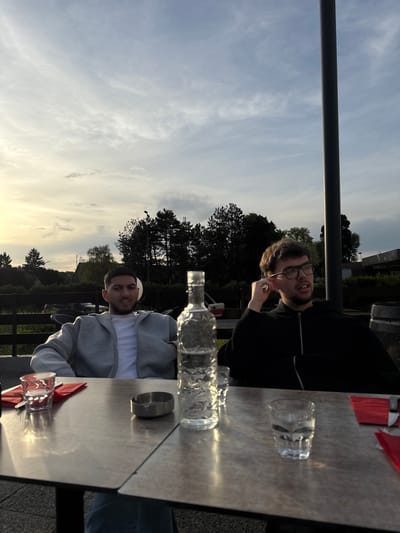 Two men are seated at a table outdoors. One man wears glasses and a black top, holding something to his ear. The other man wears a grey hooded sweatshirt. A bottle and glasses are on the table. The location appears to be a restaurant or cafe in Les Ulis, France, as suggested by the surroundings.