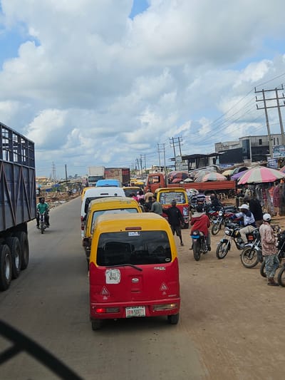 The image depicts a congested street scene in Okha, Nigeria, likely during daylight hours given the clear sky with scattered clouds. The road is filled with various vehicles, including a large blue truck on the left, several yellow and white vans, and numerous motorcycles. People are visible riding the motorcycles, some walking, and others appearing to be vendors or shoppers at roadside stalls that are set up along the right side of the road. These stalls have colorful umbrellas providing shade. The general environment appears to be an urban or semi-urban area with buildings and utility poles visible in the background. The road surface is unpaved or dirt in some areas, suggesting it might be a less developed or commercial part of the city. The overall impression is one of a busy marketplace or transit hub with significant vehicular and pedestrian activity.