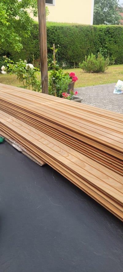 A stack of wooden planks lies on a dark, textured surface, likely a table or patio. The planks are light brown with visible wood grain and are arranged in a neat pile, angled slightly to the right. In the background, a garden scene unfolds. A green hedge forms a backdrop, with a pale yellow building visible above it. To the left, green foliage and white and pink roses are in bloom. A wooden post stands near the center of the image, supporting some of the greenery. The ground is paved with grey stone slabs. The overall impression is that of an outdoor setting in a residential area, possibly a backyard.