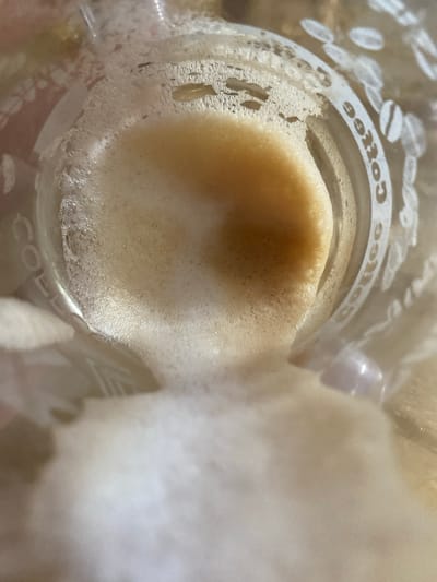 The image shows the inside of a glass with a foamy beverage, likely coffee, and the words "Coffee" printed around the rim. The beverage is a light brown color with a thick layer of foam on top. Some of the foam appears to be spilling over the side of the glass. The glass is transparent and shows reflections of light. There are also some coffee beans depicted in white around the rim of the glass. The image is taken from a top-down perspective, looking into the glass. There is no indication of the location being in the city of Maykop, Russia.