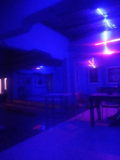 The image depicts the interior of a dimly lit establishment, possibly a bar or restaurant, in Sabon Gari, Nassarawa, Nigeria. The scene is dominated by vibrant blue and purple neon lighting, casting a dramatic glow on the surroundings.
In the foreground, a table with chairs is visible, suggesting a seating area for patrons. The furniture appears to be simple and functional. The floor is a deep blue, reflecting the ambient light.
In the mid-ground, a raised platform with steps is present, leading to a darker area with what appear to be framed artworks or decorative pieces on the wall. A single window with bright white light emanates from it, possibly an entrance or another room.
The background features a ceiling with a unique, textured design, further illuminated by the neon lights. Several neon light fixtures are suspended from the ceiling, some forming geometric shapes and others appearing as straight lines of light. One fixture prominently displays a pinkish-red hue intersecting with white and blue lights.
The overall impression is that of a modern, atmospheric venue designed for entertainment or social gathering. The low lighting and intense colors create a specific mood, potentially for nightlife or a themed event. No individuals are clearly identifiable in the scene, though shadows and indistinct shapes hint at the presence of people.