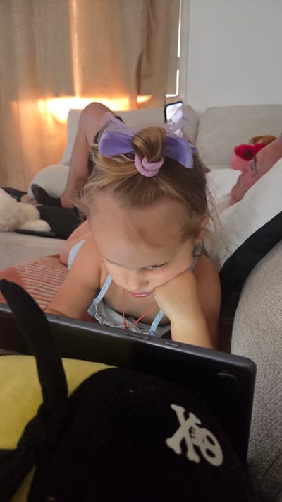This image depicts a young child, possibly a girl, sitting on a couch. She has blonde hair styled in a bun with a purple bow and a pink scrunchie. She is wearing a light blue denim dress. Her right hand is holding a red rubber band stretched from her fingers to her chin. She appears to be looking at a tablet device, which is partially visible in the foreground. The tablet is black and has a skull and crossbones design on a black accessory placed next to it. The background shows a couch and a window with sunlight streaming through, suggesting it is daytime. The scene appears to be indoors. There is no visible text in the image. The overall impression is of a child engaged in an activity with a tablet.