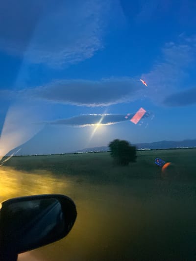The image appears to be taken from inside a moving vehicle at dusk or night, with a view through the windshield. The left side of the image shows a reflection of the sky with light clouds, and possibly a bird or small aircraft. In the center of the windshield, there is a reflection of an orange sign. Outside the vehicle, there is a field or grassy area, with a tree visible near the center of the image. A vehicle side mirror is visible in the lower right corner, with the car's headlights illuminating the road. There are faint lights of what could be a city in the distance and a partial moon in the sky. The image does not provide specific clues about the location in Pishtigovo, Bulgaria.
