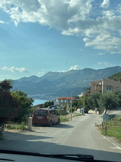 The image depicts a scenic view of a coastal area, likely in Blizikuće, Montenegro, with a road running alongside lush greenery and a dramatic mountain range in the background. The sky is a vibrant blue, dotted with fluffy white clouds, suggesting a clear and pleasant day. Sunlight streams down, casting shadows on the asphalt road, indicating it is likely daytime, possibly late morning or afternoon.
The road is bordered by trees and vegetation, with a few buildings visible, including a multi-story house with a balcony. Several vehicles are parked along the roadside, one of which is a red van with text on its side. The text on the red van reads "PUNIME NDERTIMI" which translates to "CONSTRUCTION WORKS" in Albanian, along with a license plate "AB 334FD". The presence of these buildings and vehicles suggests a populated area, perhaps a residential or commercial zone within a village or town.
Further in the distance, the shimmering blue of the sea is visible, with some structures suggesting a coastal town or resort. The majestic mountain range dominates the background, its slopes covered in dense green foliage, creating a striking contrast with the clear sky and the tranquil sea. The overall impression is one of natural beauty and a serene, Mediterranean atmosphere. No specific activity or event is clearly taking place; the scene appears to be a snapshot of everyday life in this picturesque location.